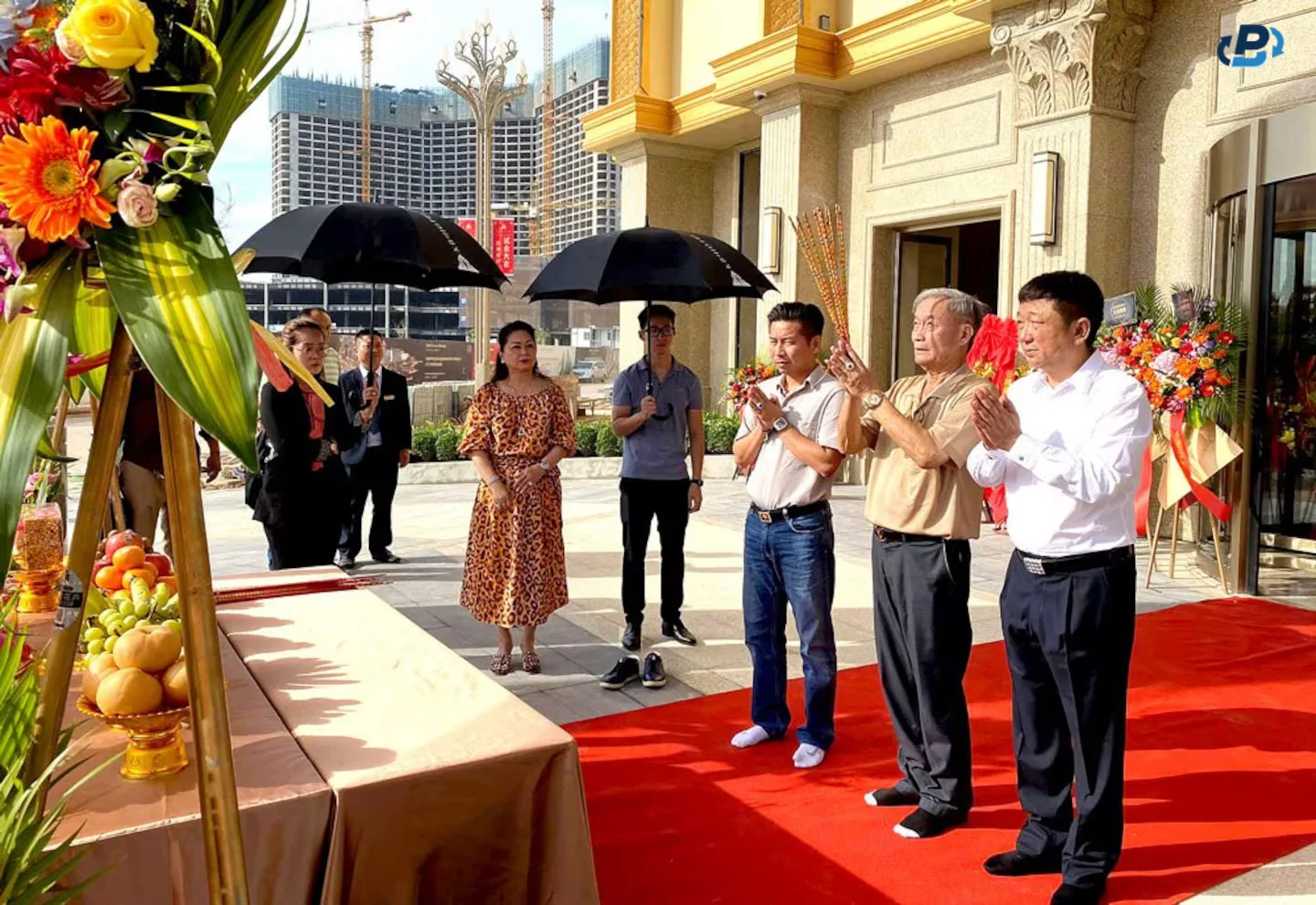 Photo showing three men standing with their hands clasped in prayer on a red carpet and their shoes off, in front of a table with flowers and bowls of fruit, and to their right people holding two black umbrellas on a sunny day