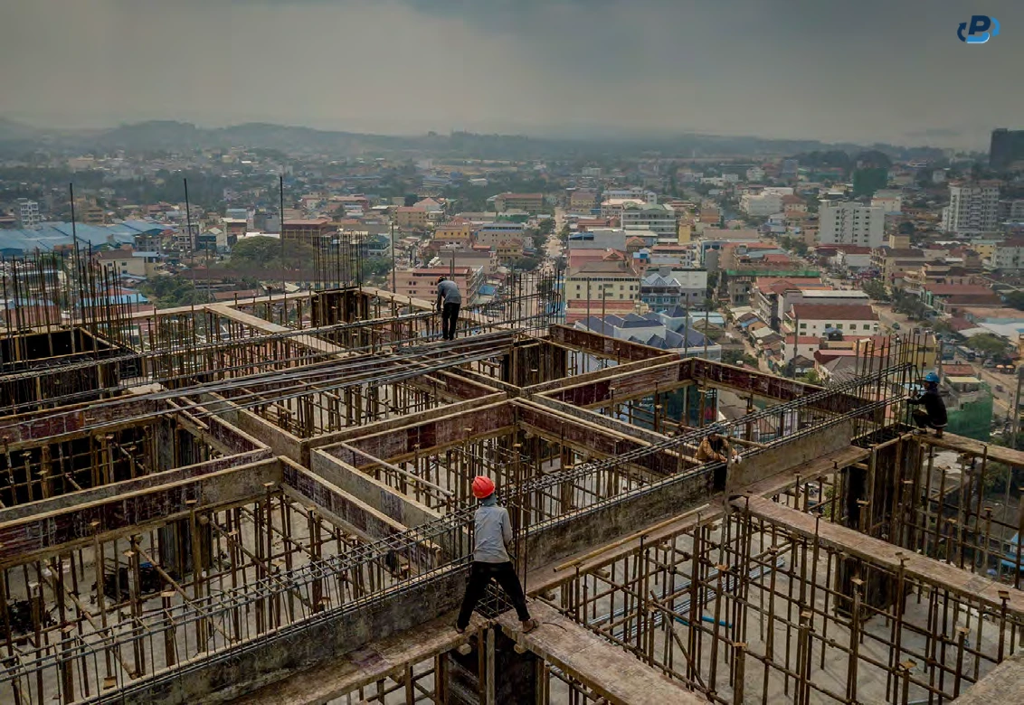 Photo showing workers in safety flip-flops installing rebar on a high-rise, and in the background we see a quaint city with low-rise buildings